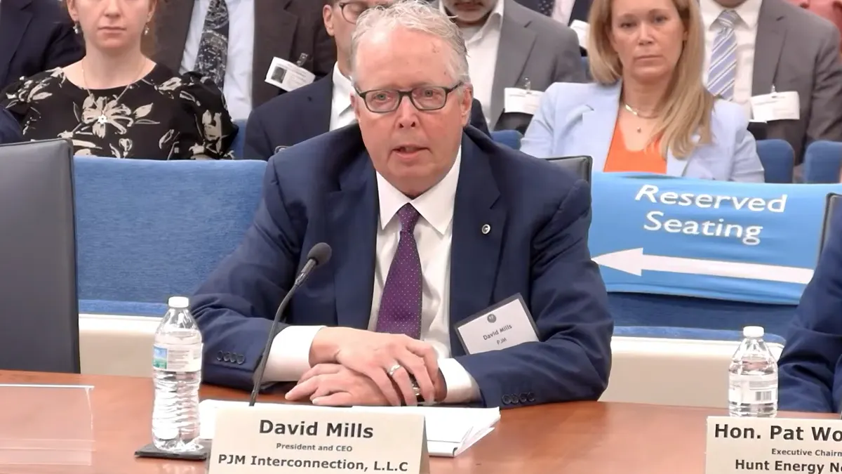 A man sits at a desk in front of a sign that says "David Mills president and CEO, PJM Interconnection."