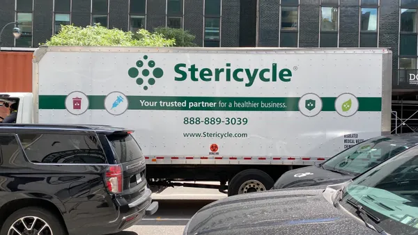 A truck with Stericycle branding sits among cars on the road with a tall office building behind it.