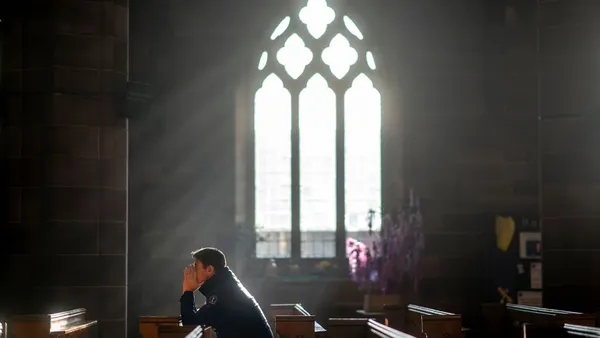 A man in a coat sits in a pew praying while light streams through a window in the background of a stone church.