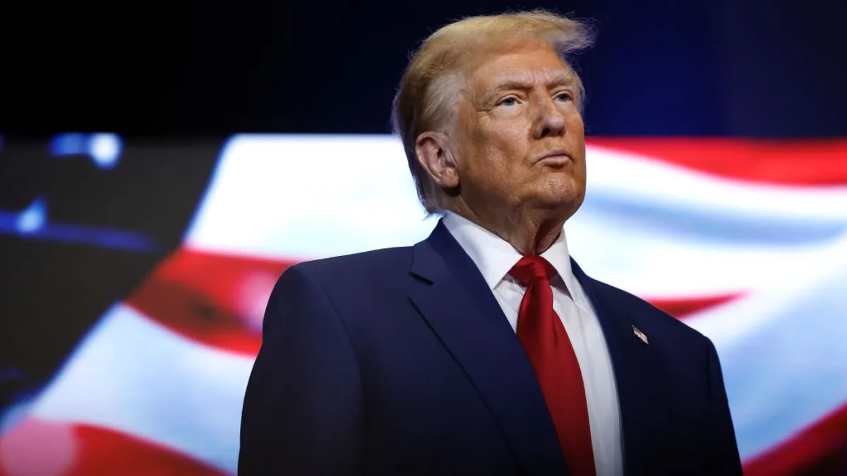 Donald Trump in business attire stands in a stage with an U.S. flag in the background