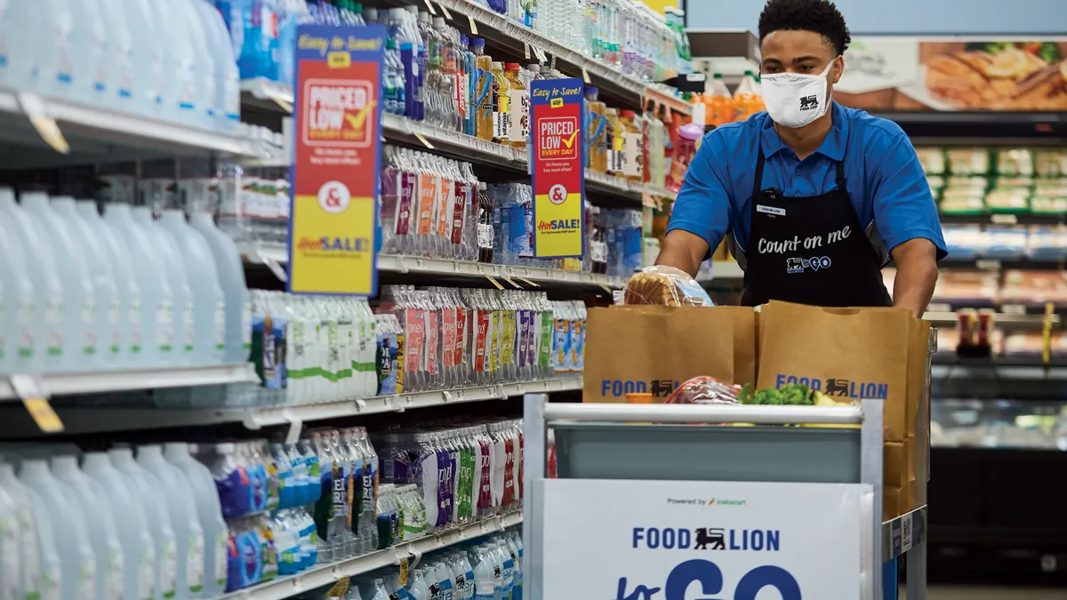 A grocery worker in a blue shirt and black apron that says "Count me" pushes a cart down filled with paper bags and food down a grocery aisle.