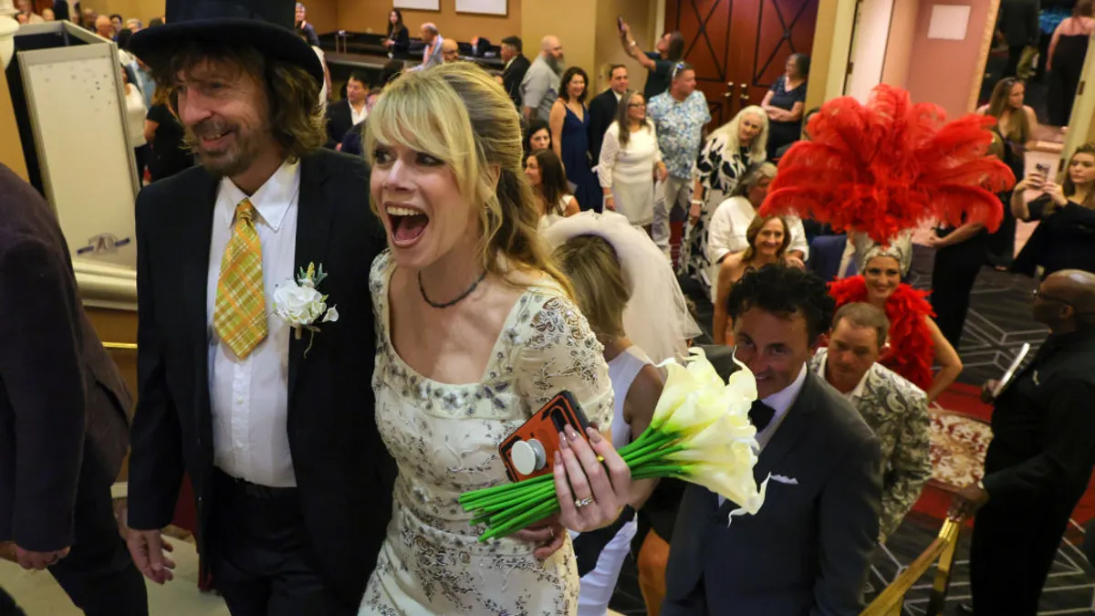 A person in a top hat and suit stands next to a person in a white dress holding flowers with a line of people behind them.