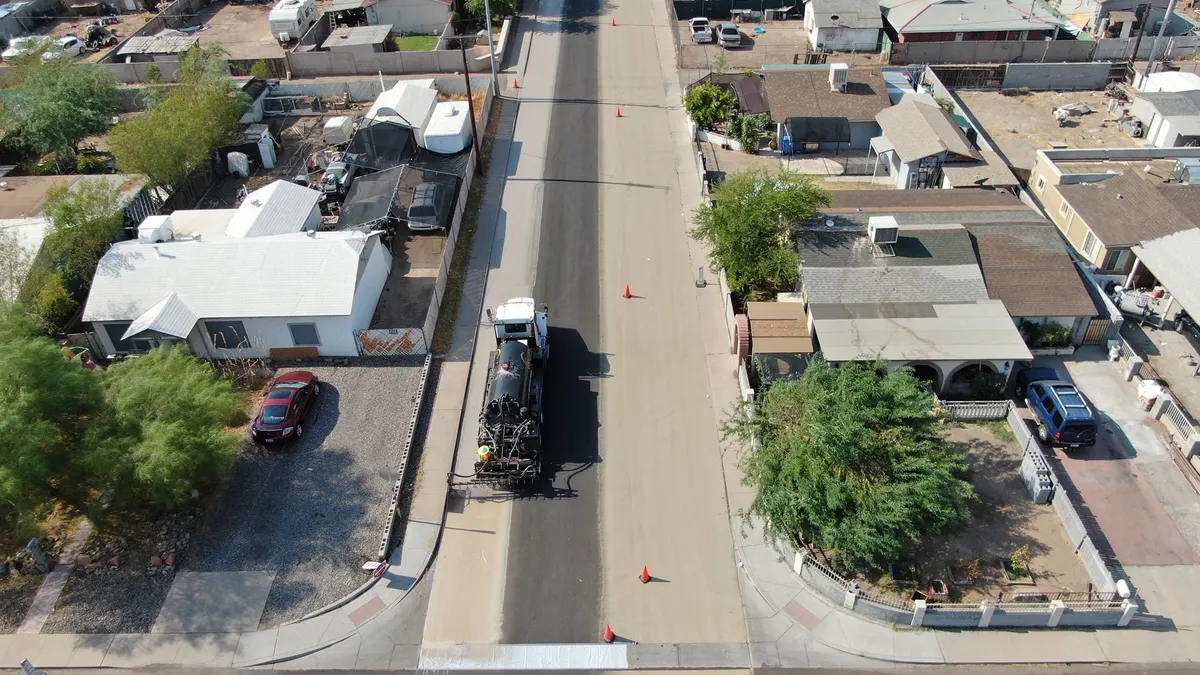 Aerial view of homes along a wide street. On the street, a truck applies a lighter-colored coating to the pavement. Traffic cones are lined up along the street.