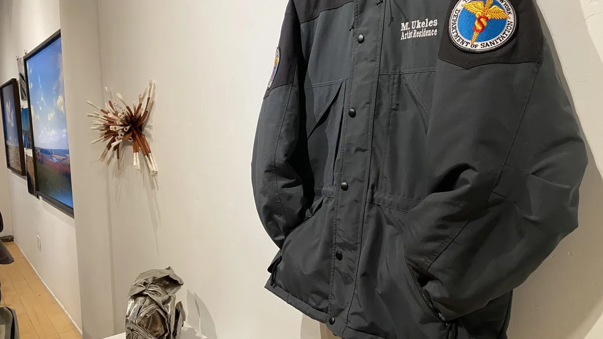 A thick jacket embroidered with a New York Sanitation Department logo and the words "M. Ukeles, Artist Residence" hangs on a gallery wall near other art pieces.