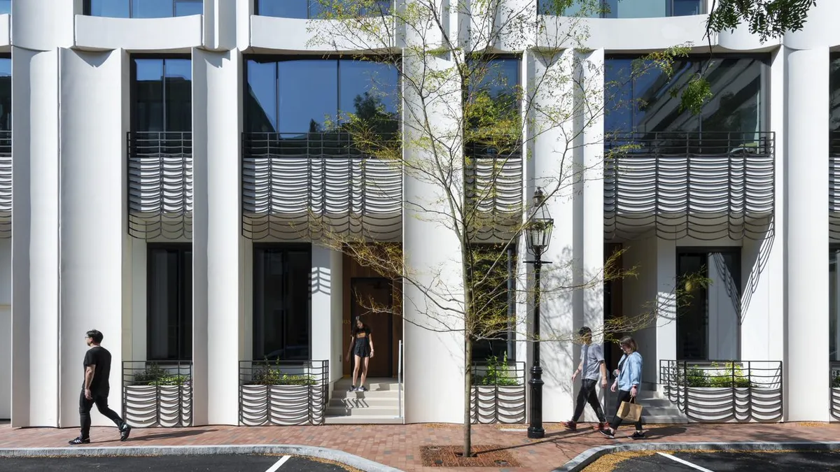 The front of a concrete building with black grate balconies. People are walking by.