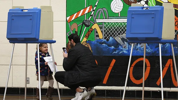 a small child is in a room with a mural and is holding a piece of paper. An adult is taking the child's photo. In front of the adult and child are voting booths