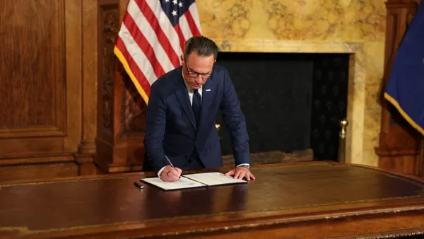 A person wearing a suit signs a paper while standing behind a wooden desk.