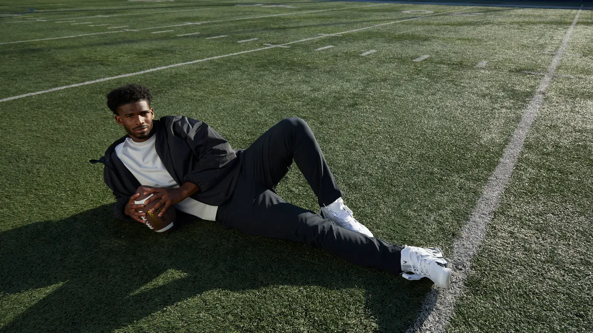 Shedeur Sanders, a football player, is pictured laying on a football field wearing a jacket and pants form Pacsun's new collection. He holds a football.