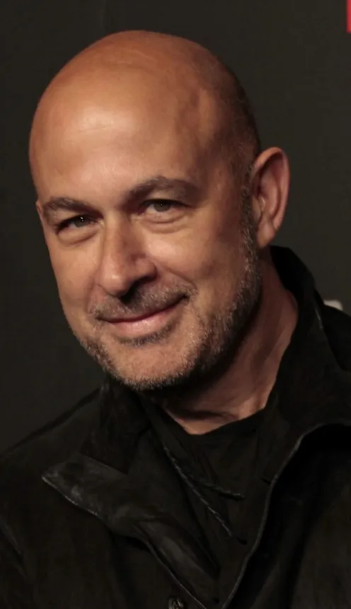 A bald person with a scruffy beard, wearing black clothing, smiles slightly.