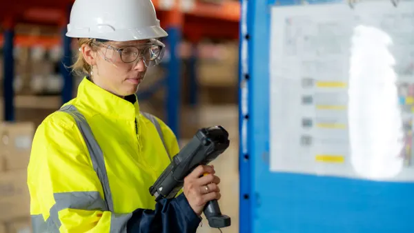 A person wearing a slate and yellow reflective jacket, white hardhat and safety goggles, looking down at a barcode scanner.