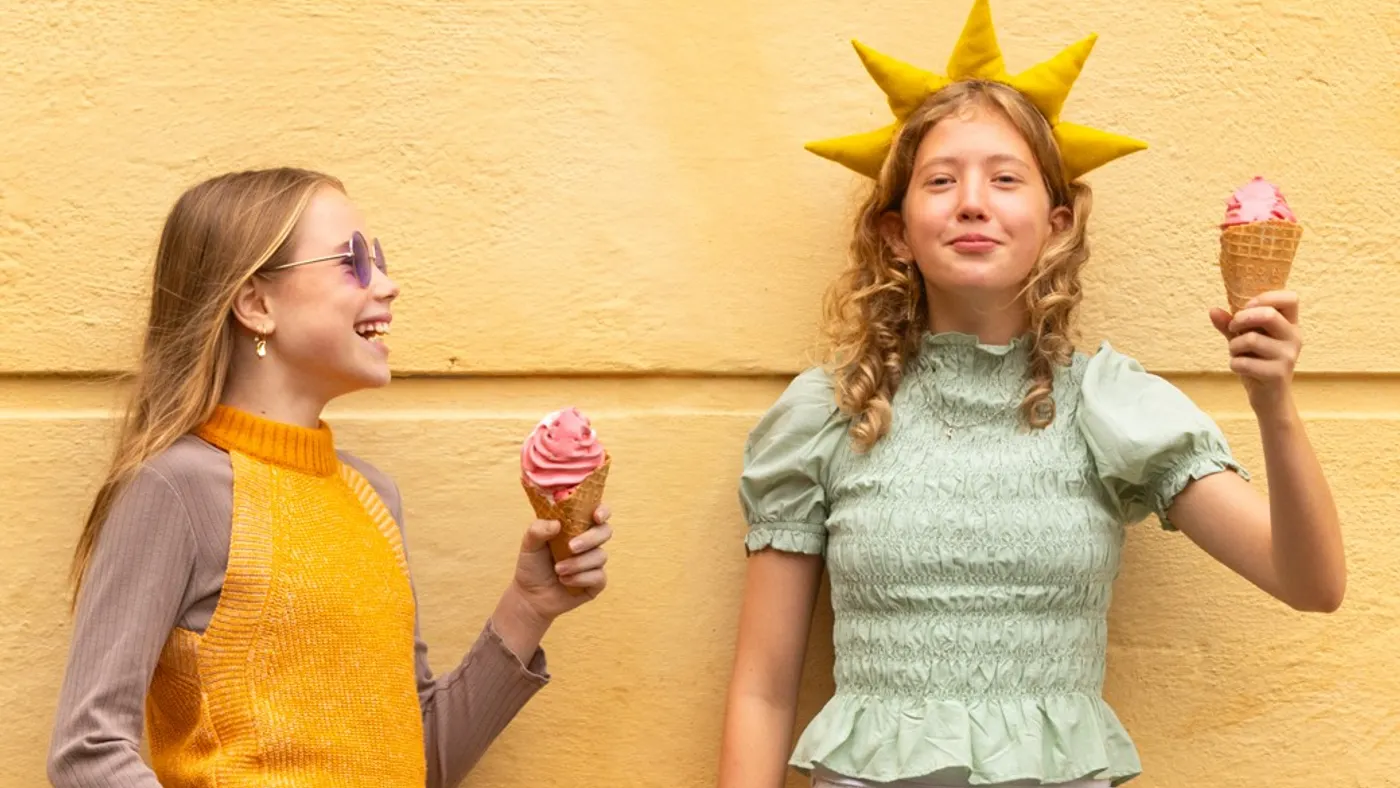 Two girls enjoying pink ice cream cones stand against a yellow wall. The girl on the left wears sunglasses and an orange sweater, smiling at her friend, who has a whimsical sun-shaped hat and a green top, creating a joyful, playful vibe.