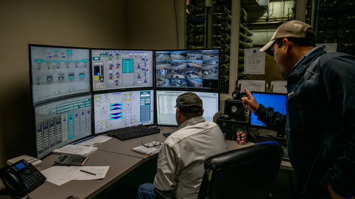 A man stands and points at a series of monitors on a desk as another man sits in front of the monitors at the desk