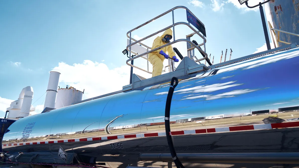 A person wearing a hazmat suit stands on top of and dips a probe into a large metal tank.