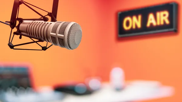 A professional podcast mic hangs in the foreground in an orange room with an "on air" sign in the background.