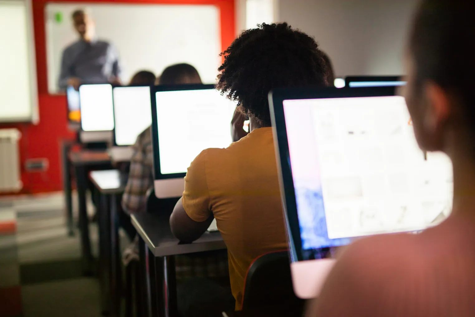 Students work on computers while a teacher instructs in front of them in a classroom.