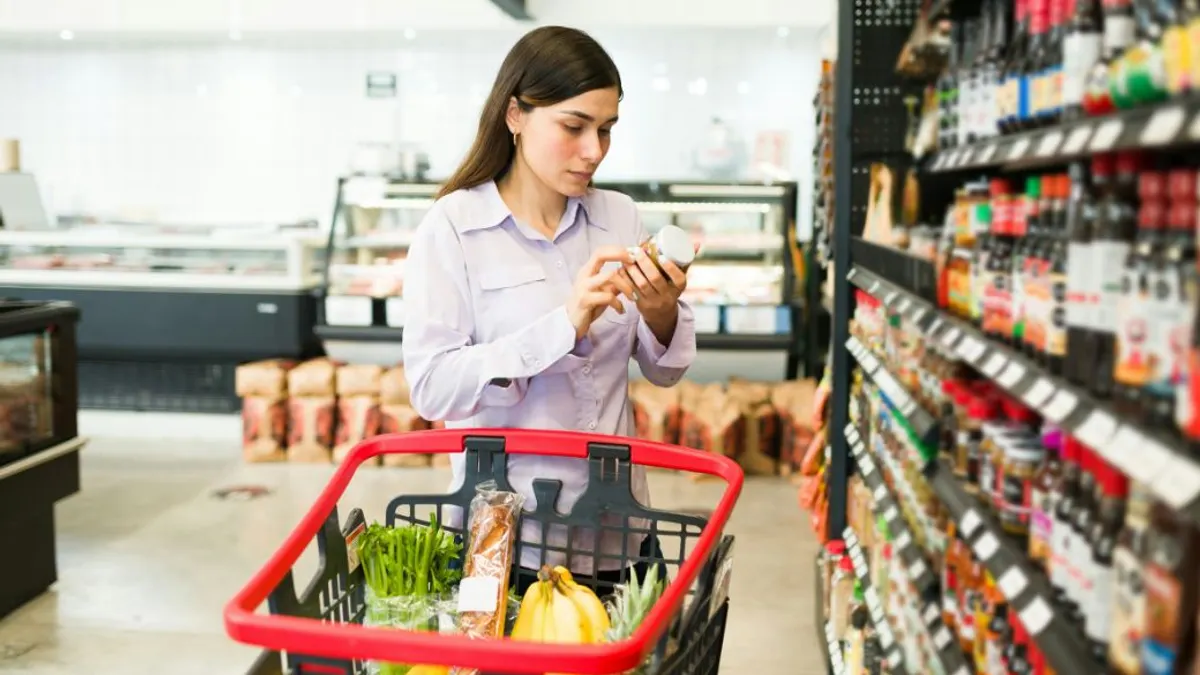 A person in a grocery store aisle examines a product container while standing beside a cart.