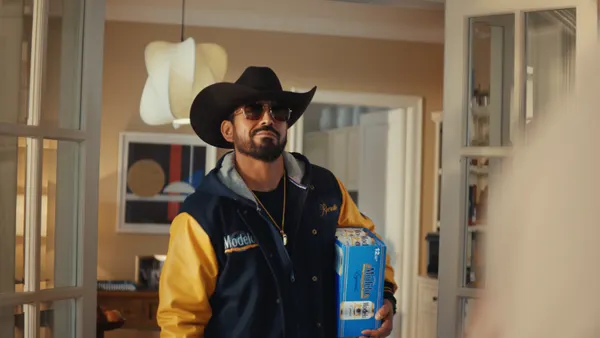 A man with a beard wearing a blue Modelo-branded varsity jacket and cowboy hat stands in an archway holding a case of beer.