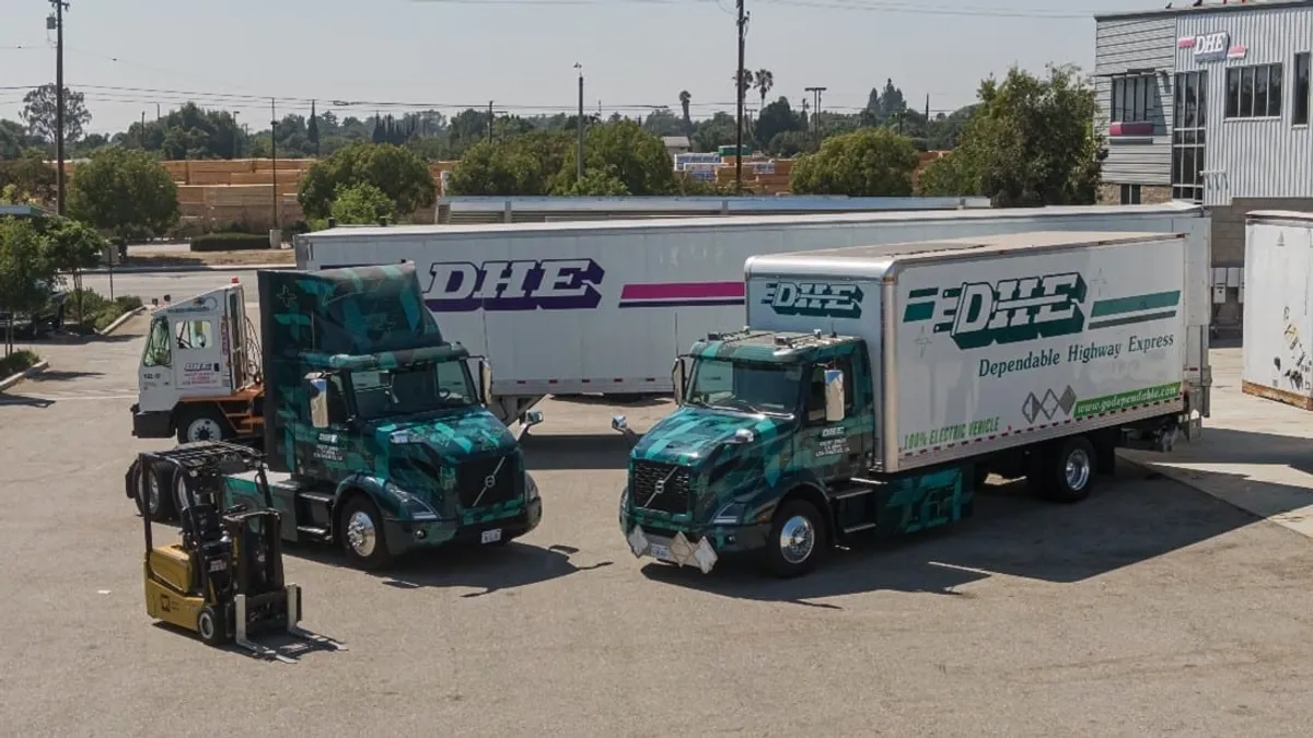 Two of Dependable Highway Express' Volvo VNR Class 8 EV trucks sit next to a forklift and another DHE truck in a parking lot.