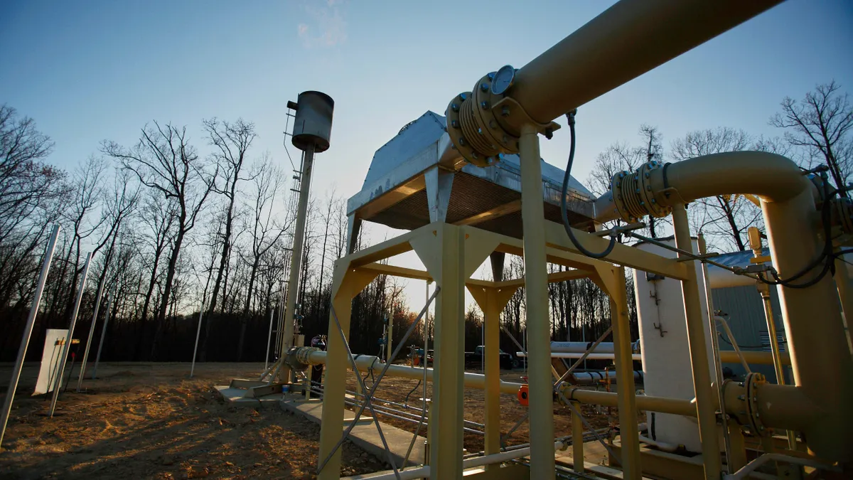 An industrial landfill site surrounded by trees is equipped with gas collection infrastructure, including vertical extraction wells and piping systems, capturing methane emissions from decomposing waste. 