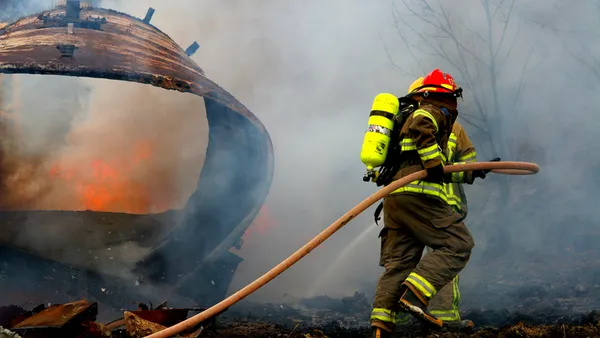 Two people in firefighting suits carry a hose outside. Fire and smoke is billowing in front of them.