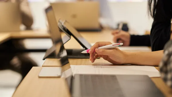 A close-up photo of an open laptop on a desk. A person's hand is near the laptop and is holding a multi-colored pen.