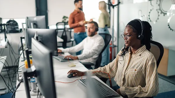 A customer service representative wearing headset works in front of a computer in a call center as her colleagues stand in the background.