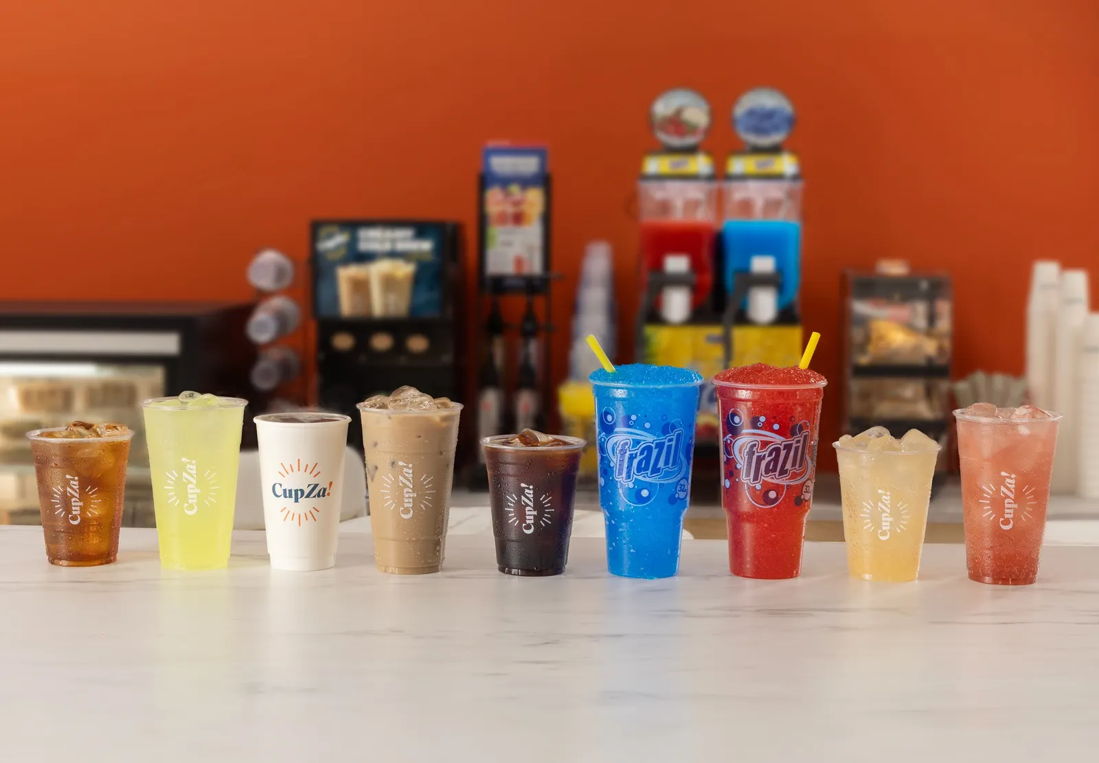 A line of cups with words like Frazil and CupZa on them containing a variety of colorful beverages sit on a white table against a background of beverage dispensers