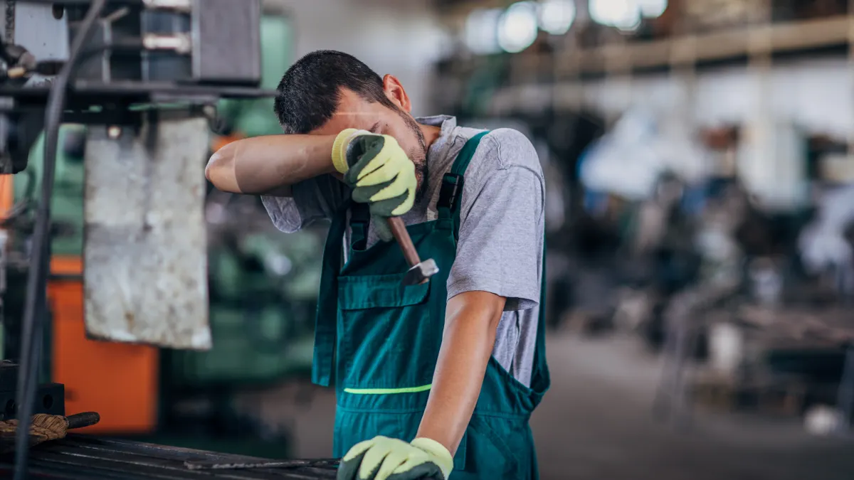 A person wearing coveralls wipes sweat from their in an industrial facility