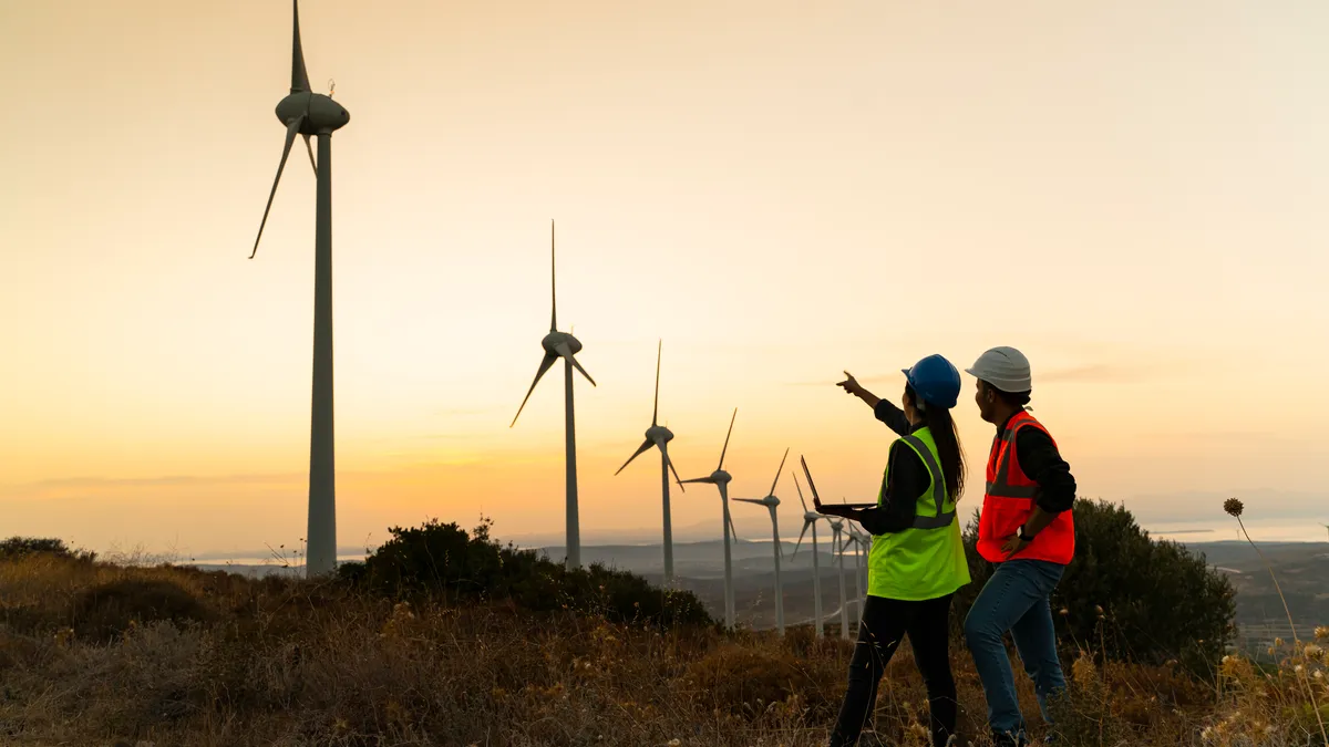 Silhouette of young engineer holding laptop computer planning and working for the energy industry and standing beside a wind turbines farm power station at sunset time