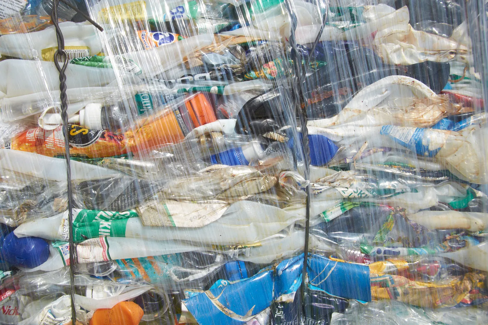 A tightly packed bale of crushed plastic bottles, compressed into a rectangular block and wrapped in clear plastic film. The bale contains various colored plastic bottles, flattened and compacted together, ready for recycling.