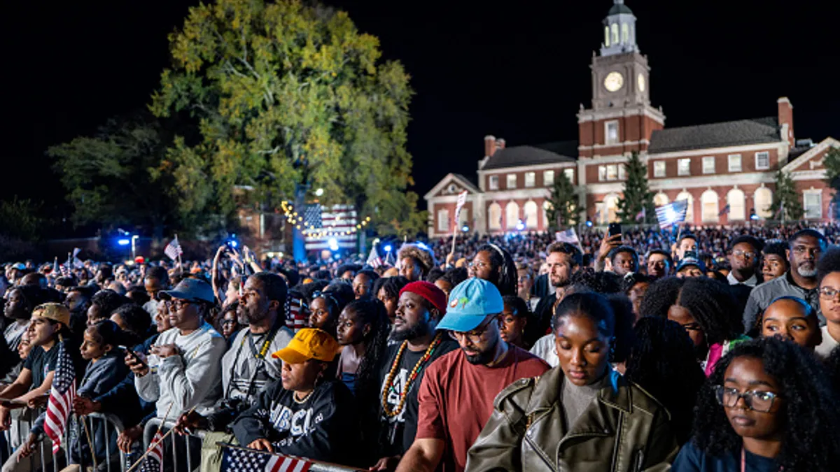 A crowd of people are standing behind a metal fence outside at night at Howard University. Some are holding American flags.