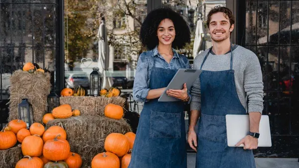 Happy young coffee shop owners waiters baristas bartenders service staff standing outdoor near their cafe with laptop and decorating pumpkins preparing for Halloween