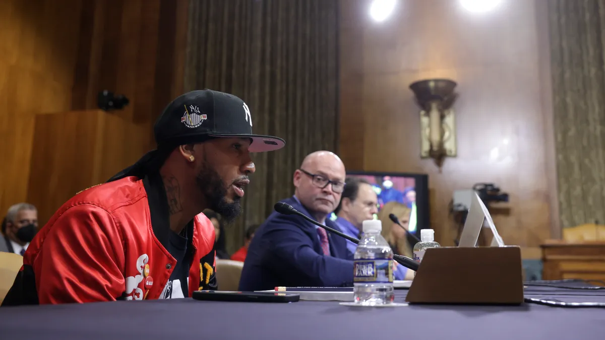 Two men in focus, sitting at a long table testifying to Congress with microphones in front of them.