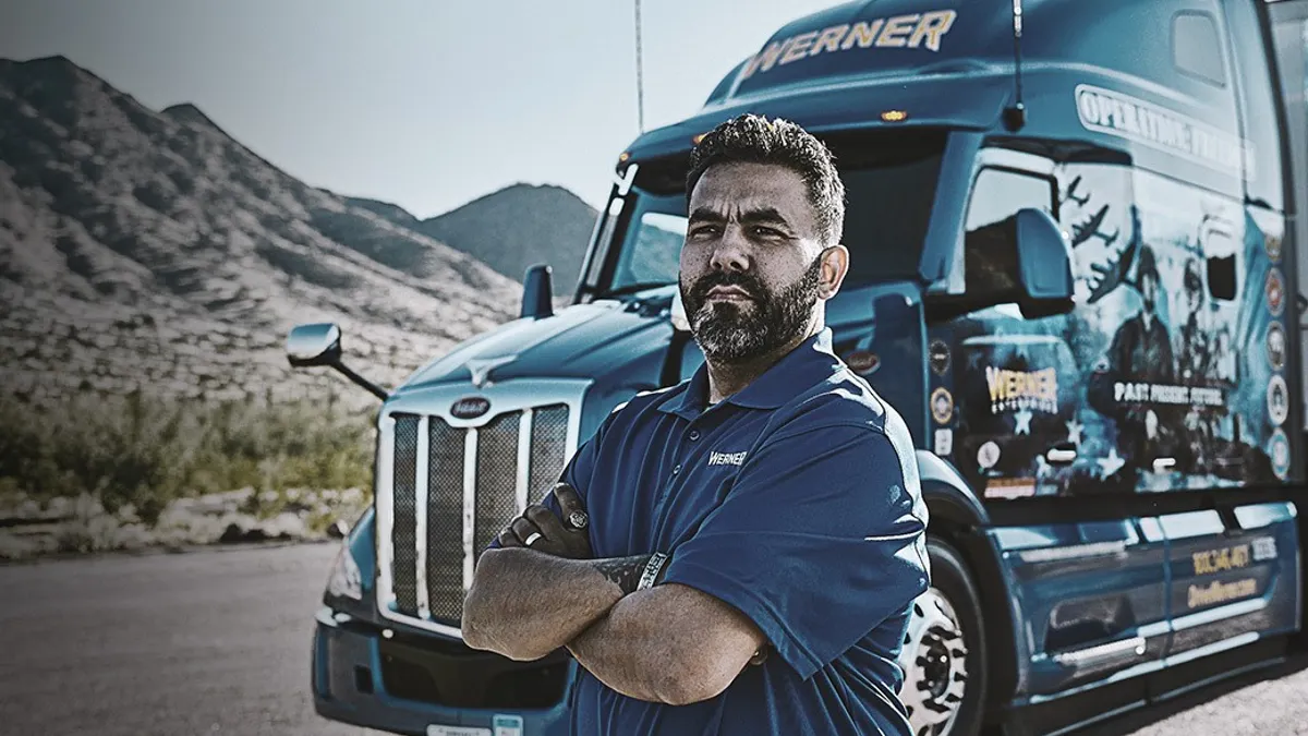 A bearded man in a blue Werner shirt stands with arms crossed in front of a decorated blue semi-truck on a mountain road.