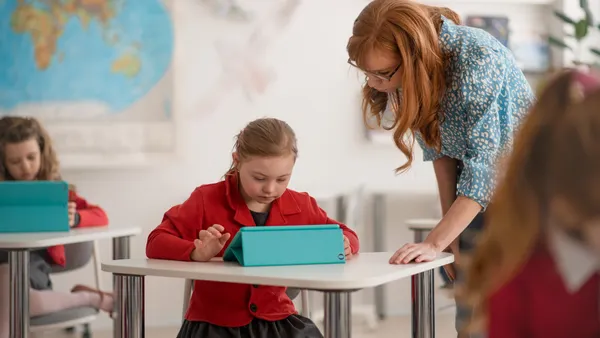 A student is sitting at a desk in a classroom and is looking at a tablet on the desk. A teacher is standing over the student.