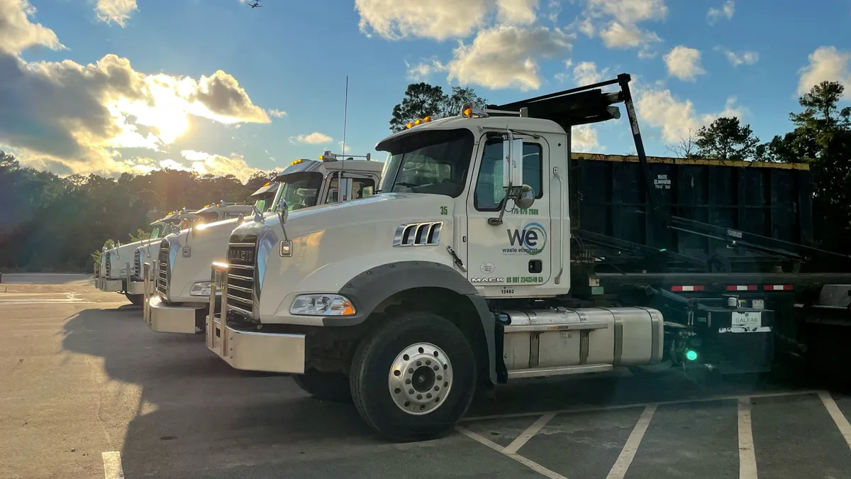 Four Waste Eliminator trucks lined up in a parking lot, with sun and clouds in the background