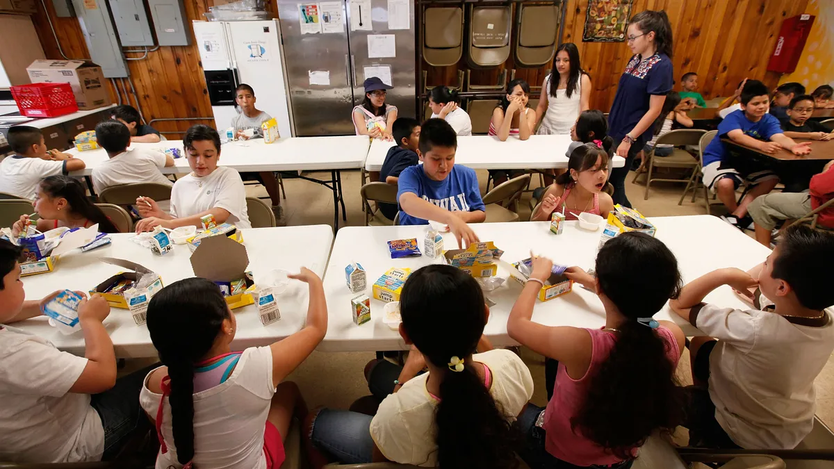 A group of children eat free breakfast together while sitting at various tables inside.