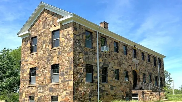 Stone hall surrounded by grass and weeds on a sunny day.
