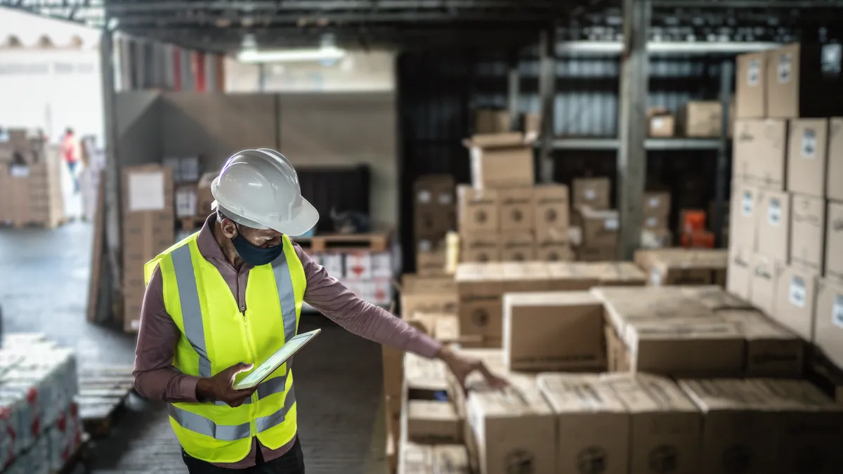 Warehouse worker wearing face mask and protective workwear checking products using digital tablet