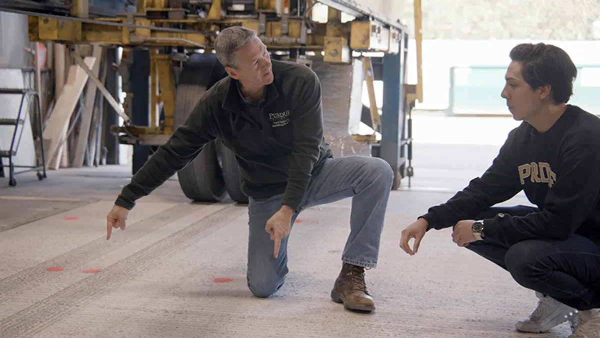 An older and younger person kneel in a workshop as they examine a grey concrete tile on the ground.