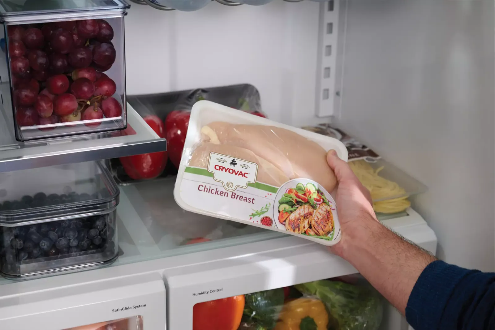 A person holding a wrapped tray of raw poultry in front of an open refrigerator with food inside.