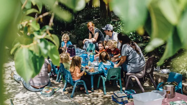 A group of children and several parents are seated around a small table outside, enjoying their lunch.