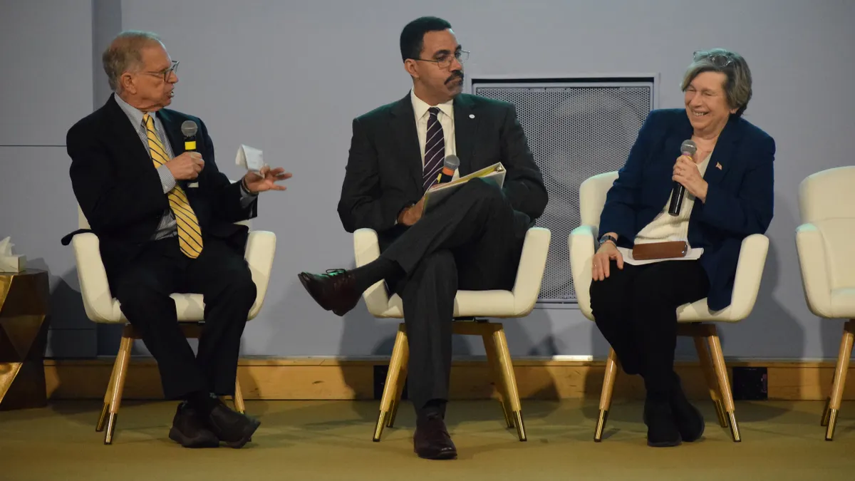 Three people are sitting in chairs on a stage. One person has a microphone