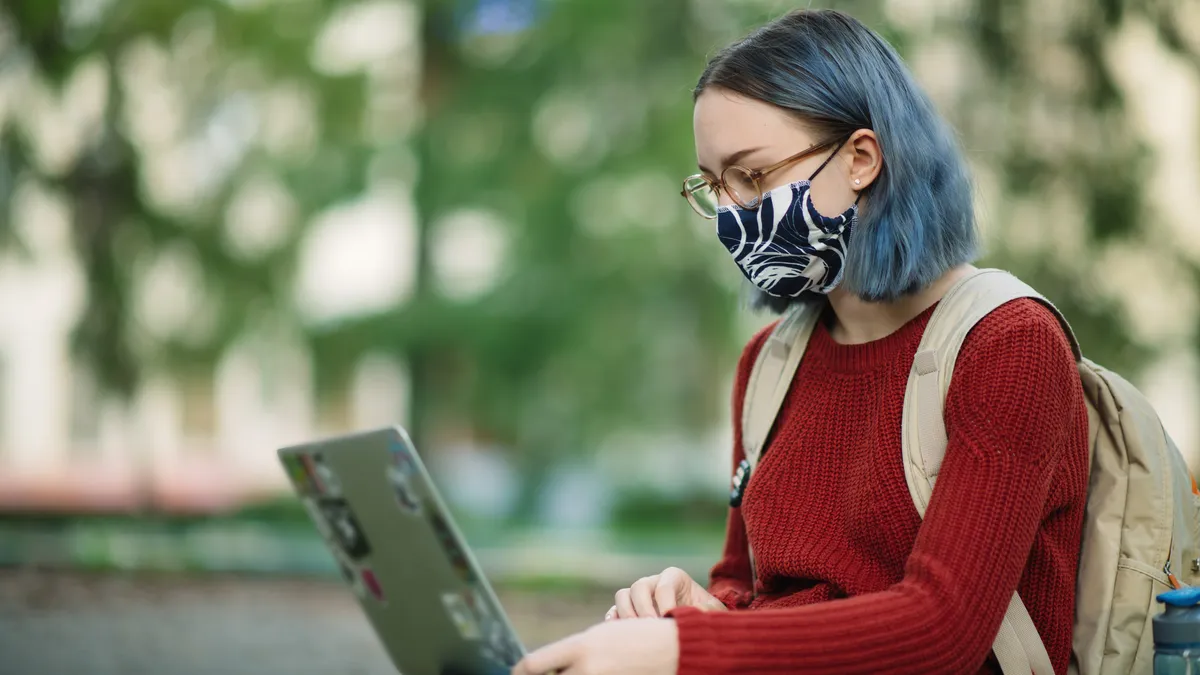 Young woman in a reusable protective face mask uses her laptop outdoors.