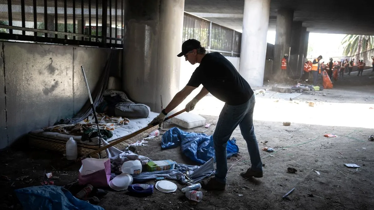 A person wearing a hat uses a tool to clean up debris and garbage under a freeway.