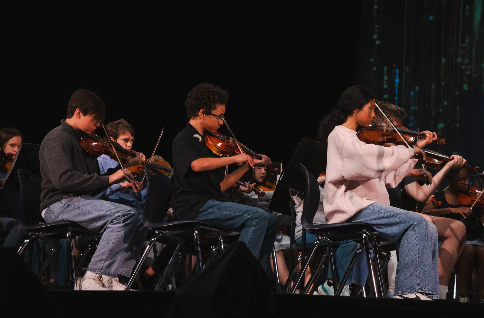 A group of students is sitting in chairs on a stage. Each are playing a violin.