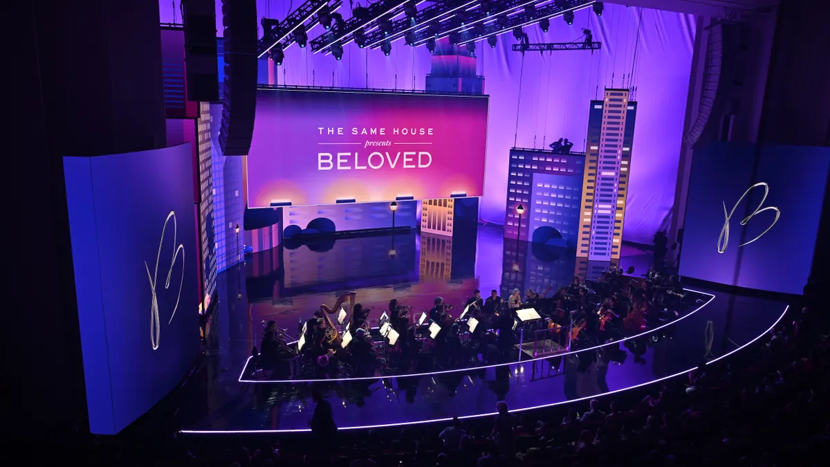 Orchestra on stage with purple lighting, large screen that says "Same House Presents Beloved."