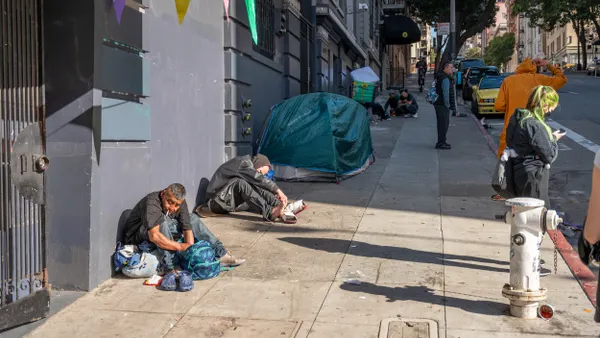 Two people sitting on a sidewalk in front of tents on a sidewalk.