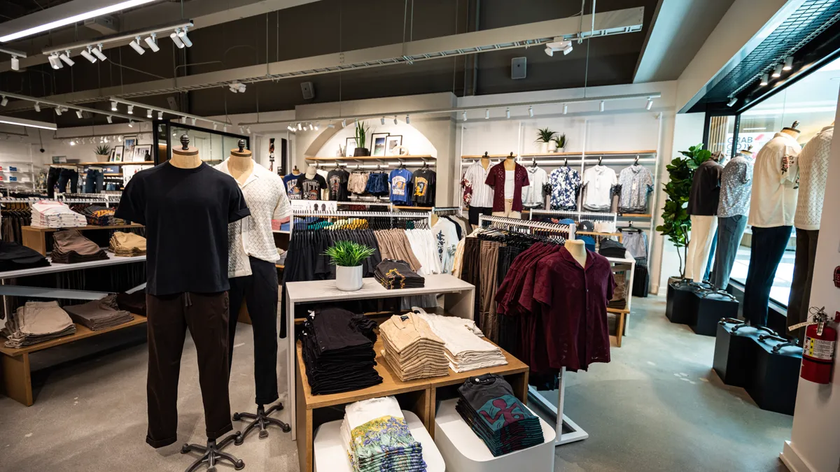 Interior shot of an Abercrombie store. Two mannequins wearing short sleeves and long pants are central, with tables and racks of clothing around them. Four mannequins stand in the main window.