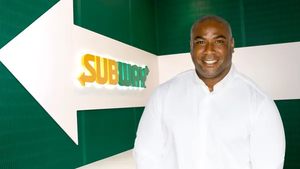 A photograph of a person wearing a long-sleeved white shirt standing in front of a green background with a Subway logo.